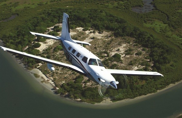 High-angle aerial view of a white Piper PA-46 aircraft banking above a circular land mass, surrounded by the shoreline and green vegetation.