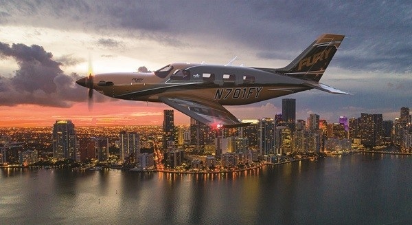 Aerial view of a Piper M700 Fury turboprop aircraft, tail number N701FY, soaring over a cityscape and water at sunset.