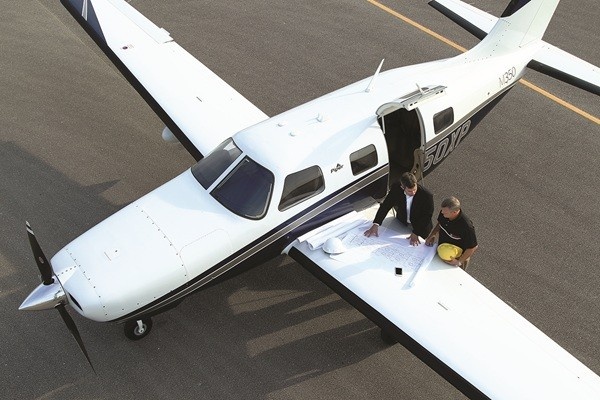 High-angle top view of a white Piper M350 aircraft parked on the tarmac with its cabin door open, as two men review paperwork spread out on the wing.