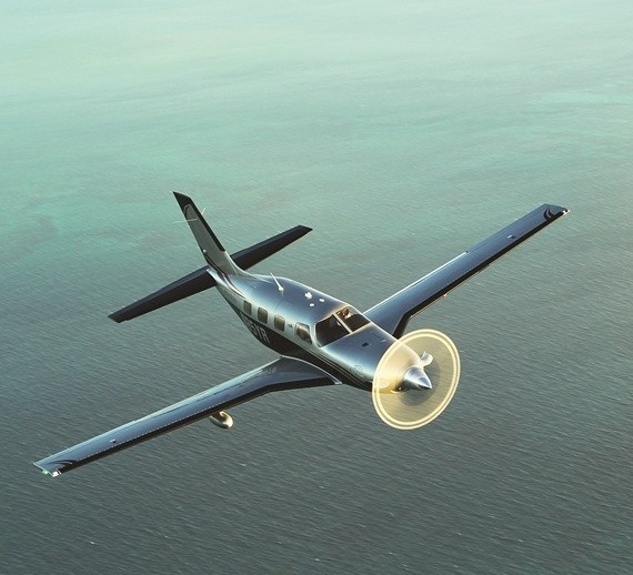 Aerial view of a single-engine turboprop Piper PA-46 Malibu Meridian aircraft in flight over green water with light reflecting off its spinning propeller.