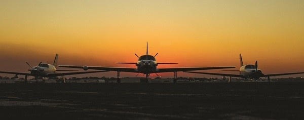 Front-facing silhouette of three Piper airplanes parked on a runway during a vibrant orange and yellow sunset.