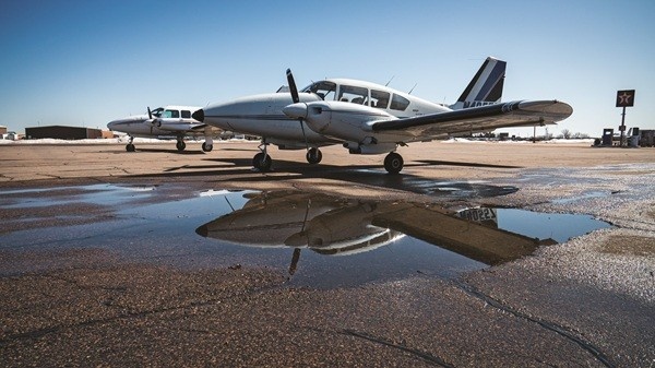 A white Piper PA-23-250 Aztec twin-engine aircraft parked on partially wet pavement with its reflection shown in the water.
