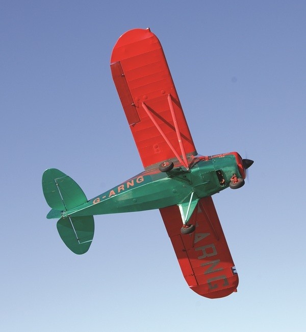 Underside view of a vintage green and red Piper PA-22-108 Colt aircraft, modified with tailwheel landing gear and tail number G-ARNG, as it flies in clear blue skies.