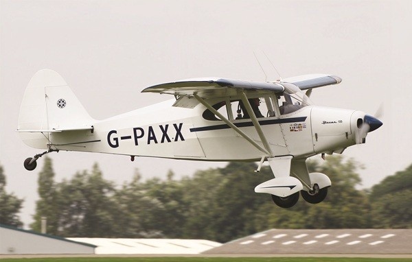 White Piper PA-20 Pacer aircraft with tail number G-PAXX during takeoff, with blurred green trees and hangar rooftops in the background.