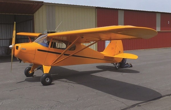 Bright yellow vintage Piper PA-15 Vagabond aircraft parked on an asphalt tarmac in front of airport hangars.