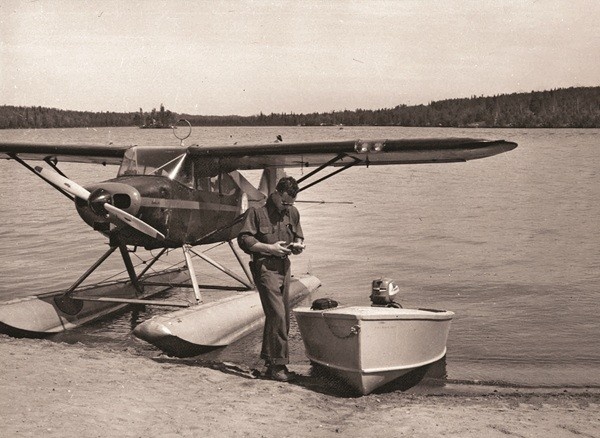 Vintage black and white image of a Piper PA-20 Pacer floatplane parked on a sandy shoreline, with a man standing next to a small motorboat, against a background of a large lake and tree-covered hills.