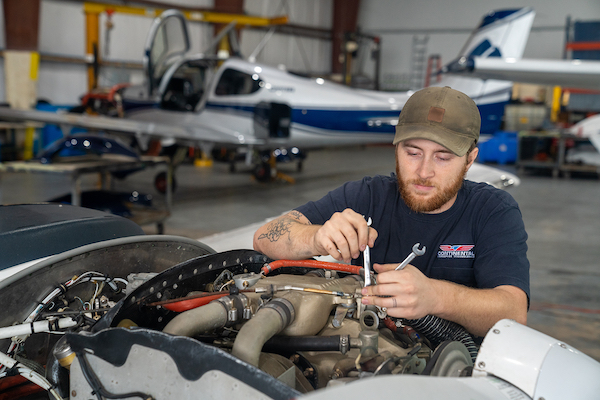 A mechanic in a baseball cap adjusts a Continental aircraft engine using two wrenches inside a large workspace, with a small plane in the background.