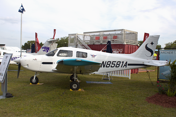 Piper Aircraft Celebrates First Pilot 100i Delivery Outside Continental U.S. to Inter American University A white Piper PA-28-181 Pilot 100i showing tail number N8581A is parked on green grass with a tall booth and flag in the background.