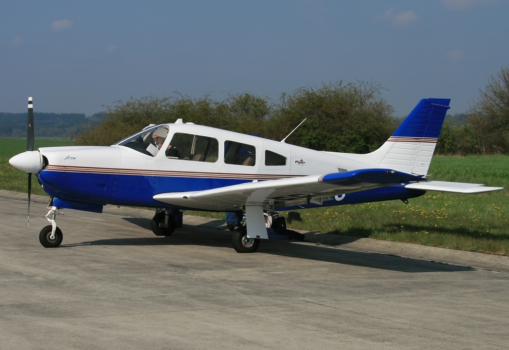FAA Revises and Clarifies the Cherokee Wing Spar Proposed Airworthiness Directive 2018-CE-049-AD A white and blue single-engine Piper PA-28R-201 Arrow light aircraft parked on a tarmac with green grass and shrubs in the background under a blue sky.
