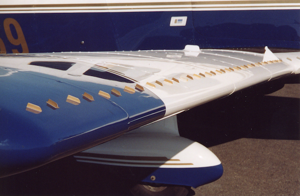Row of small metal vortex generators installed along the upper leading edge of a white and blue aircraft wing.