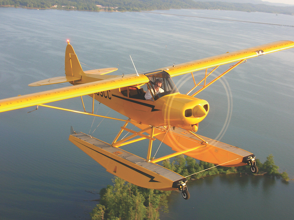 Yellow Piper Super Cub floatplane equipped with a float landing gear flying low over a calm body of water with green islands.