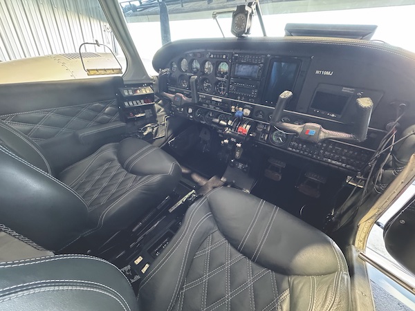 Detailed interior view of a Piper aircraft cockpit showing the flight controls, digital avionics dashboard instrument panel, and premium black leather pilot seats with white diamond pattern stitching.