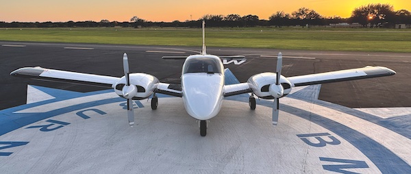 Wide front-facing view of a white twin-engine propeller Piper aircraft sitting centered on a blue and white painted airport ramp during sunset.