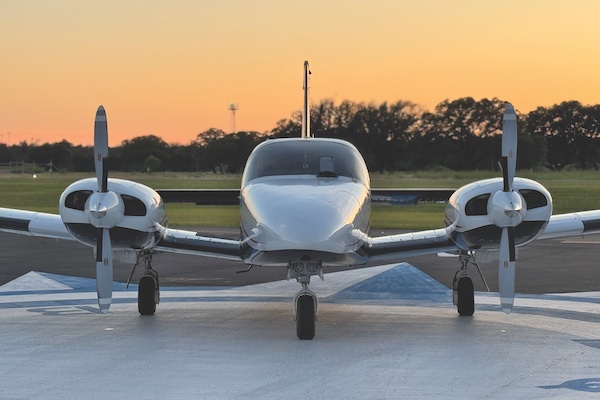 Head-on, front-facing view of a white twin-engine propeller Piper aircraft parked on a concrete ramp under a warm, glowing sky at dusk.