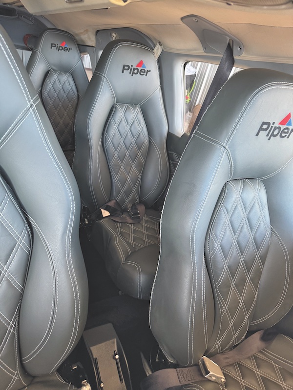 Interior cabin view of a Piper aircraft showing premium grey leather seats with white diamond stitching and the "Piper" logo embroidered on the headrests.
