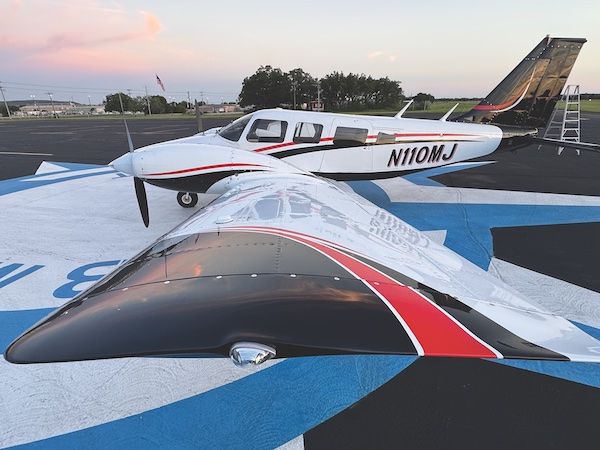 Close-up, elevated view looking down the wingtip of a white Piper Seneca V twin-engine airplane with black and red accent stripes, showing the registration tail number N110MJ parked on a blue and white painted airport ramp.