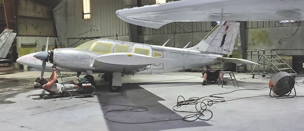 Mechanics working on a stripped-down twin-engine Piper aircraft inside a maintenance hangar with its windows taped for painting.