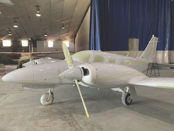 Side view of a white twin-engine propeller Piper aircraft with masked windows and taped propeller blades inside a paint hangar bay.