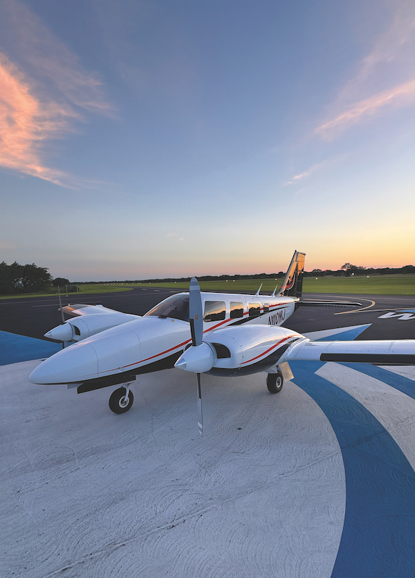 Vertical view of a white twin-engine propeller Piper aircraft parked on a painted runway ramp under a soft dusk sky with wispy clouds.