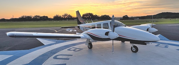 Wide-angle panoramic shot of a white twin-engine propeller Piper aircraft parked on a painted airport ramp at sunset.