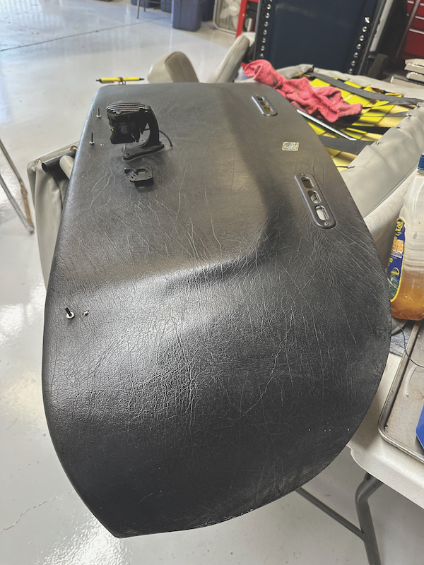 A detached, black textured aircraft Ashby glareshield panel resting on a table in a workshop hangar, showing small top-mounted components, such as a compass mount and defrost vents.