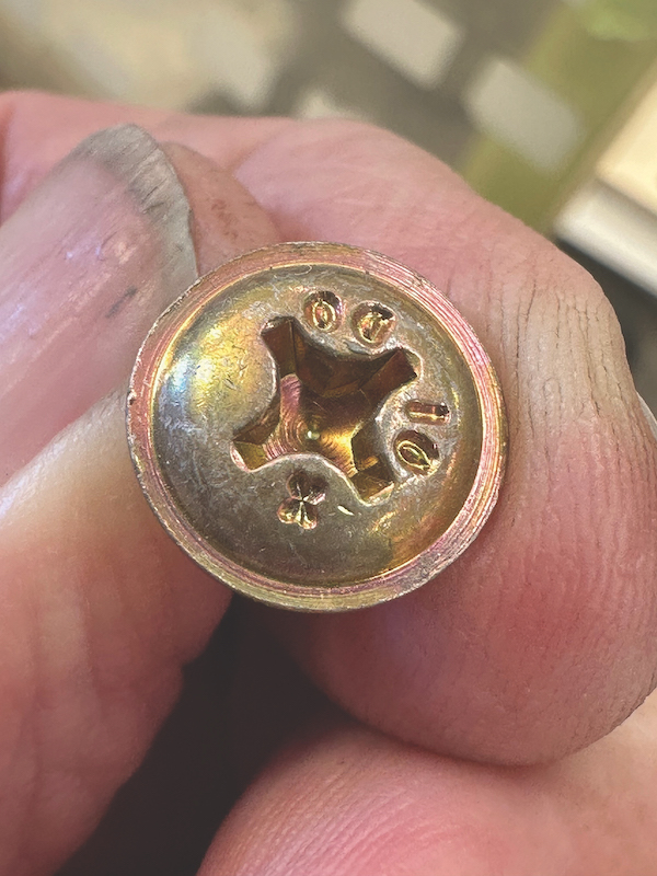 Close-up of a brass-colored AN525 structural screw held between two fingers, showing the cross-shaped Phillips head and distinct structural identification markings stamped on the surface.